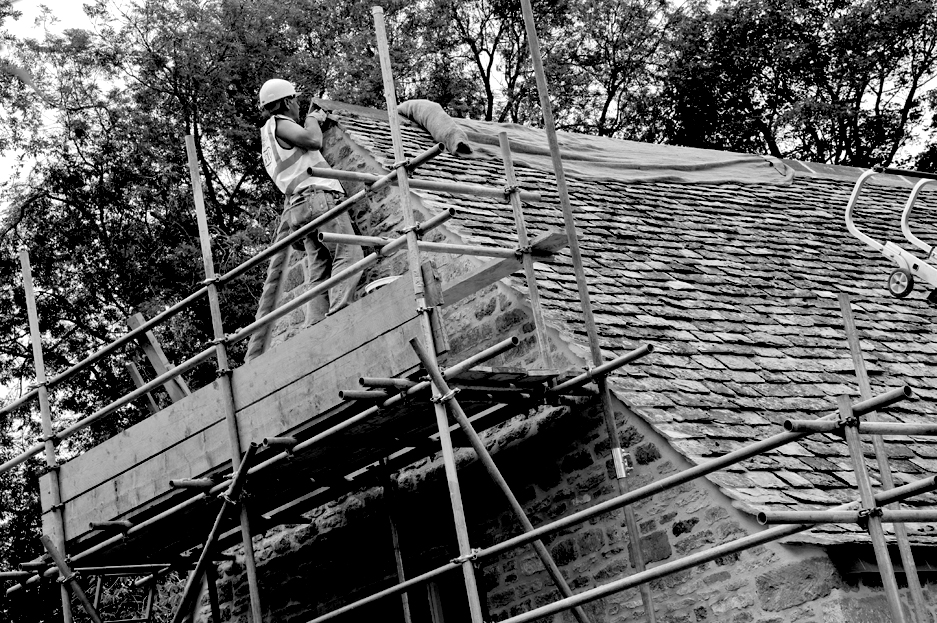 A craftsman up a scaffold tower working on the roof of a large country cottage.